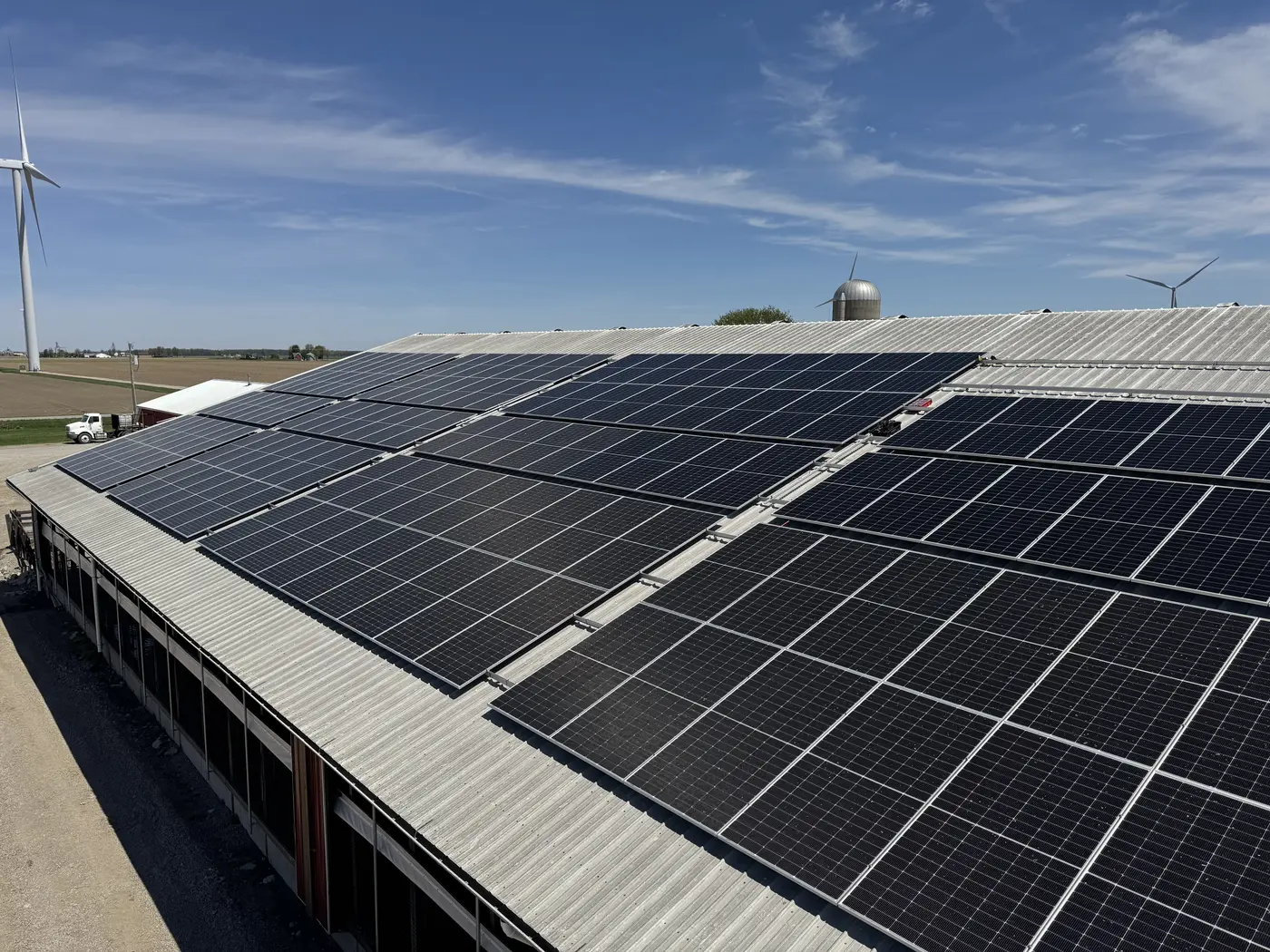 Rooftop solar panels on a dairy barn at TRT Sinn Dairy, with a silo and wind turbines in the distance